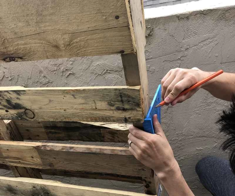 一個人用直角尺和鉛筆在木托盤上畫一條直線，用來製作書架。