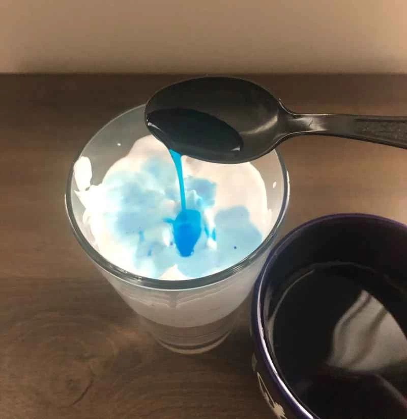 一張照片顯示,一個人正在用湯匙將有色水加入裝滿水和刮鬍膏的玻璃杯中,這是科學實驗項目的一部分,目的是在玻璃杯中製造一場「風暴」。