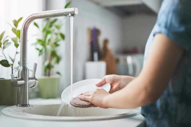 在陽光明媚的廚房裡，一個人正在流水下清洗碗碟，強調了清潔和環保的習慣。盆栽植物與清新整潔的環境相得益彰。